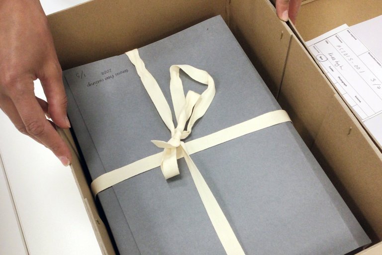 Photograph of hands opening archive box with grey archive folder with archival ribbon wrapped around. 