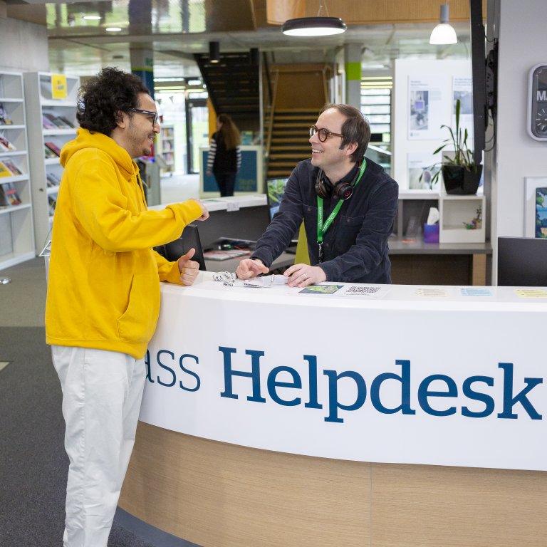 student in yellow sweatshirt speaking to staff at library helpdesk